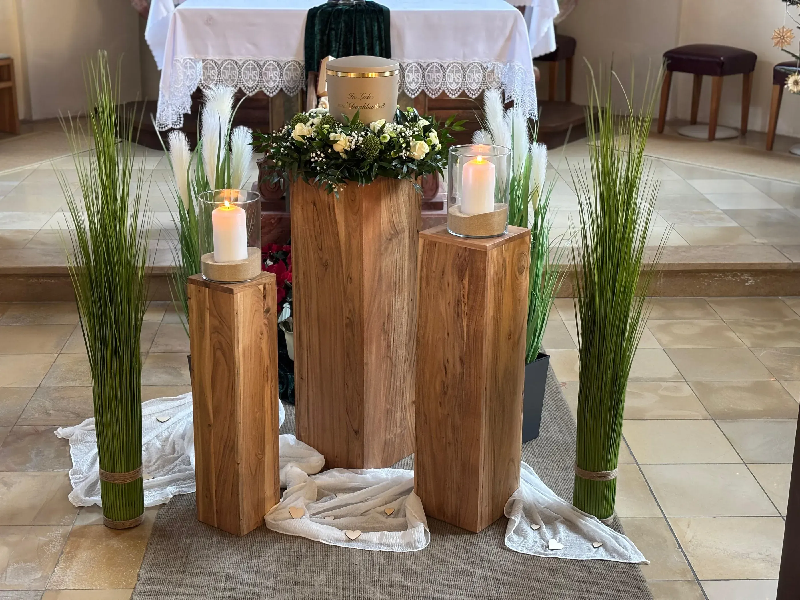 Eine Urne auf einer Holzsäule, umgeben von weißen Blumen, Kerzen und Gräsern – eine von uns gestaltete Trauerfeier